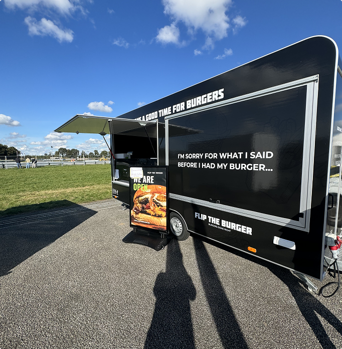 A black food truck from 'Flip The Burger' parked on a sunny day with a blue sky and scattered clouds. The truck has the slogan 'I’m sorry for what I said before I had my burger...' printed on the side. A standing signboard in front of the truck displays burger images and a message saying 'We are open'. The truck is set up near a grassy field with a few people visible in the background.